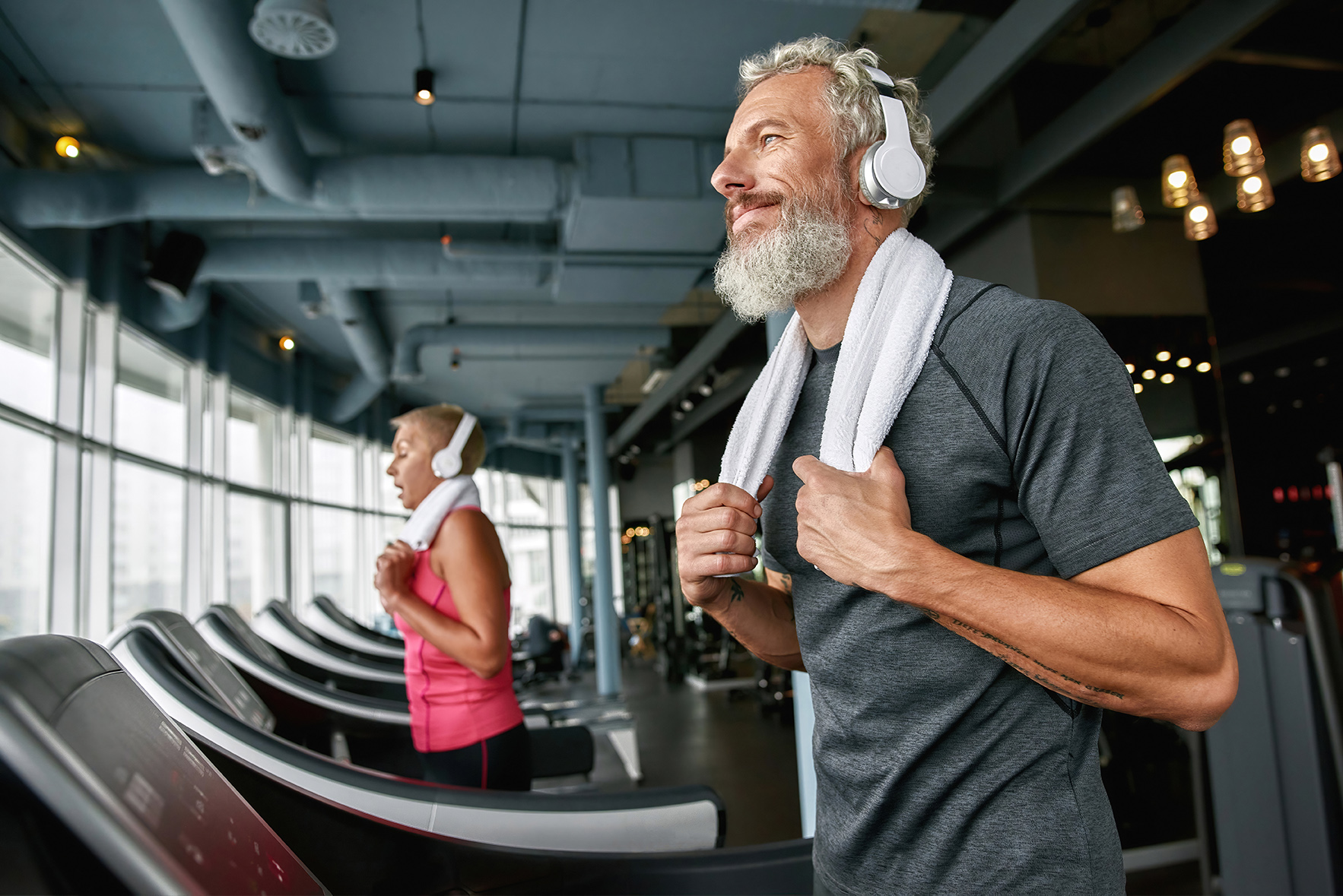 Portrait of cheerful senior man in gray t-shirt and white headphones with towel on shoulders, exercising on treadmill in gym. Healthy lifestyle concept.